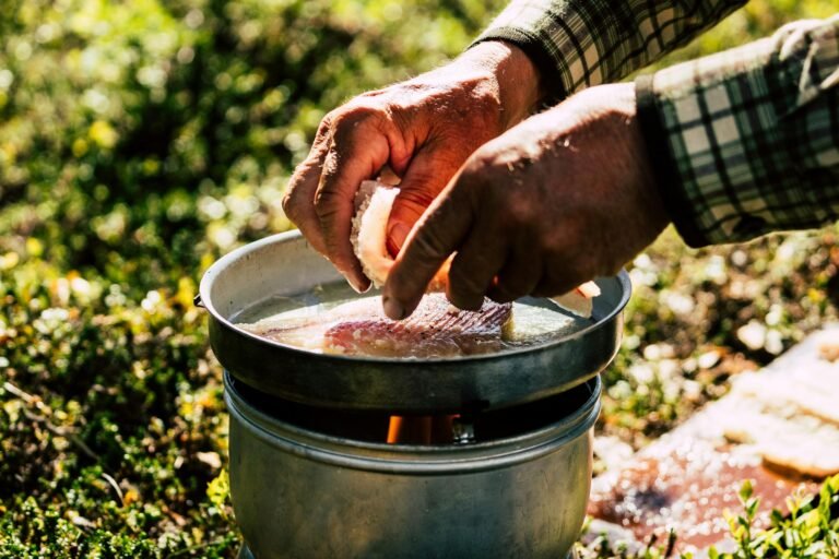 Close-up of hands cooking fish outdoors on a rustic stove, highlighting a natural and organic lifestyle.